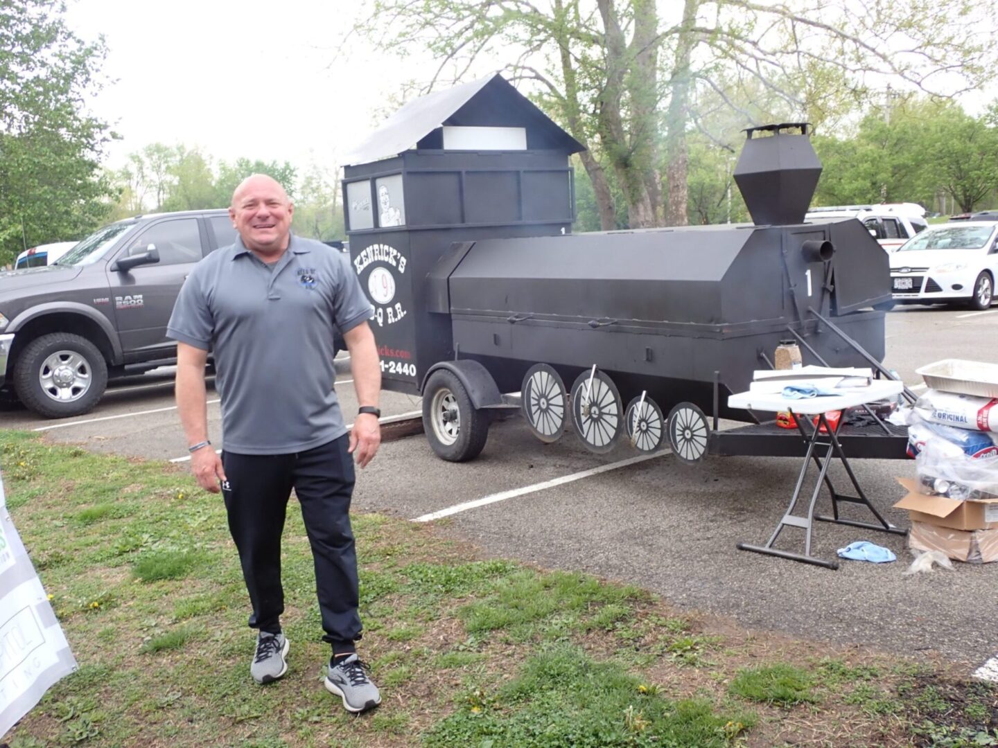Man smiling near a large, black, train-shaped grill outdoors.