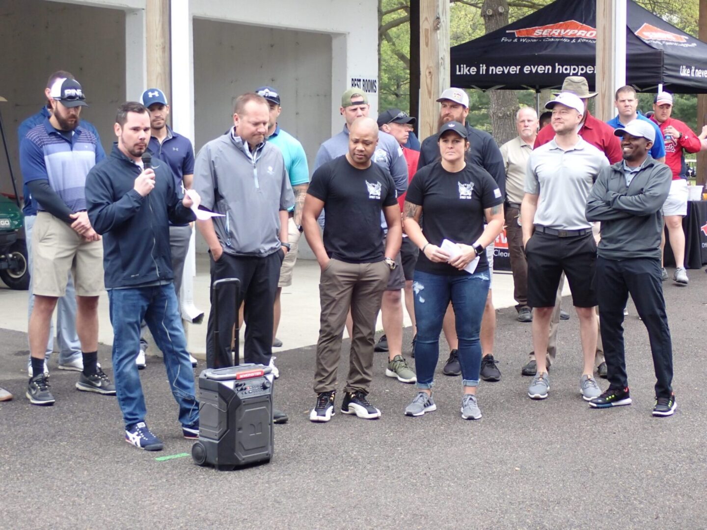 A group of men standing together, some holding papers, around a small speaker outdoors.