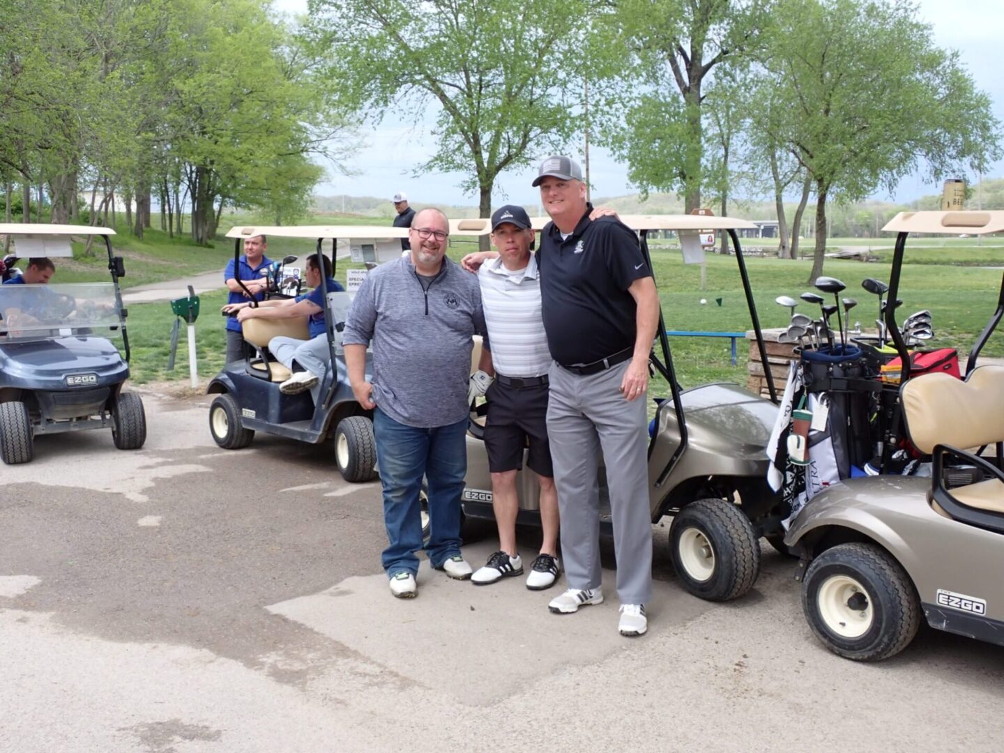 Three men standing next to golf carts on a sunny golf course.