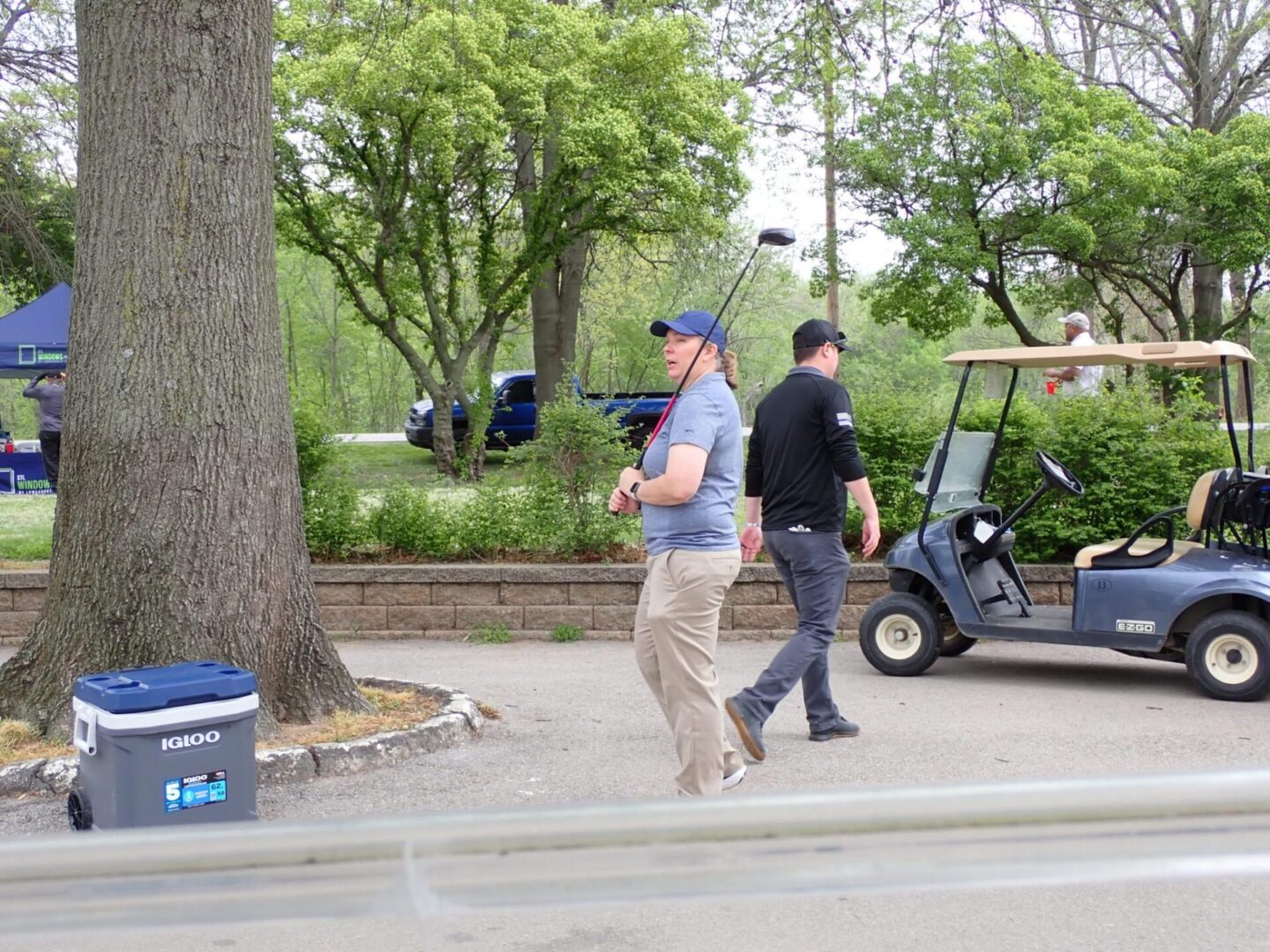 Two men walking near a golf cart on a tree-lined path.