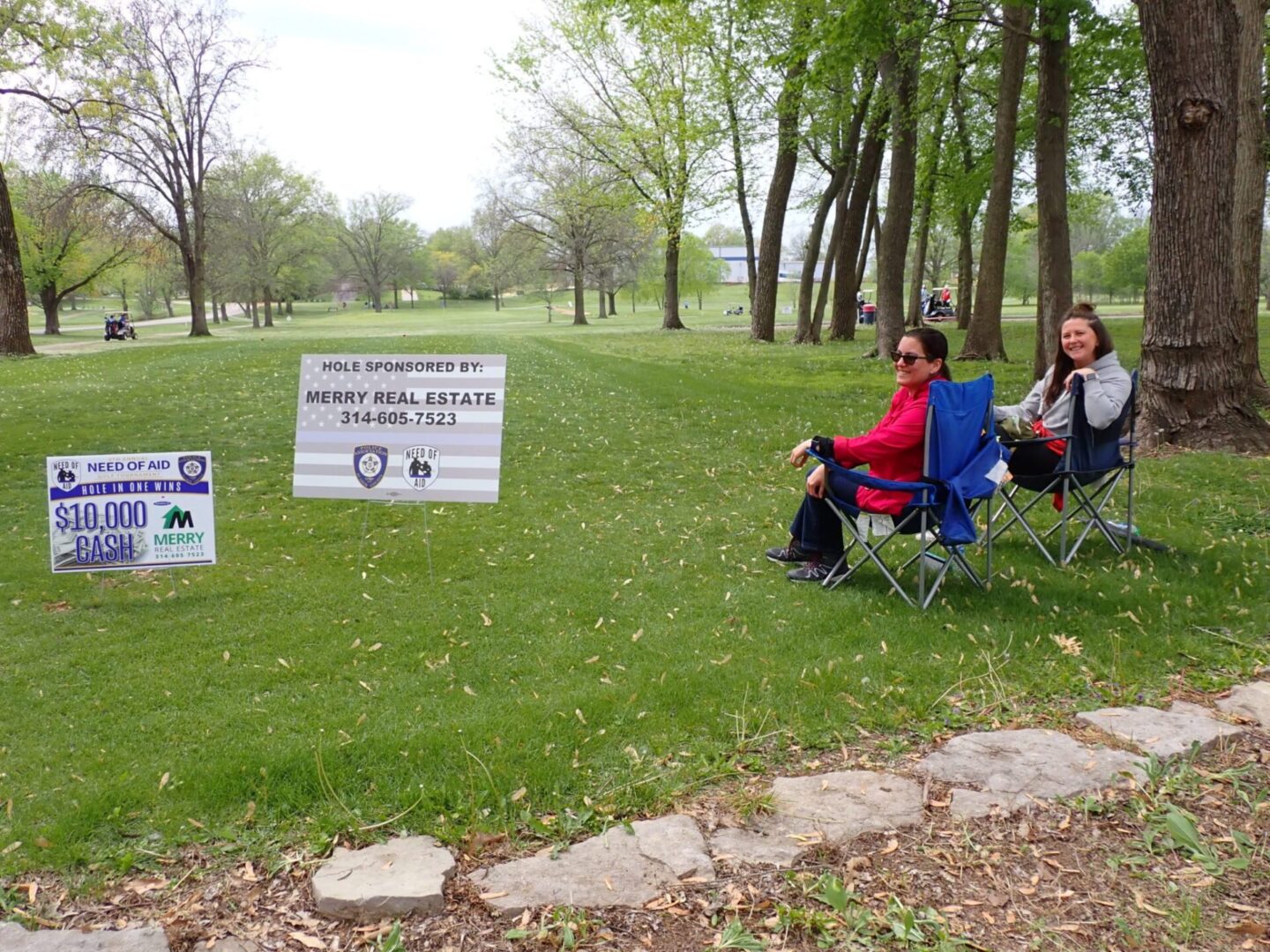 Two women sitting on chairs on a grassy field with signs around them.