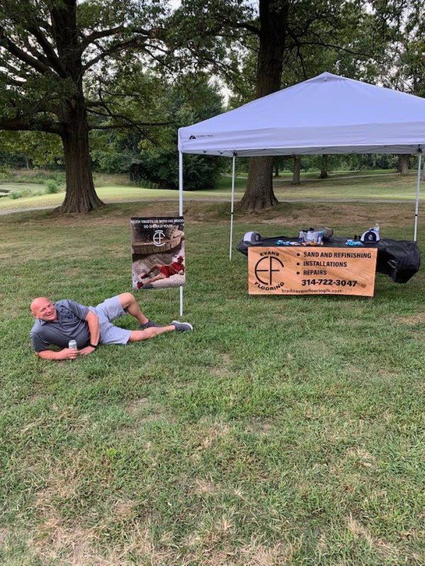 Man resting on grass near a tent with a promotional sign in a park.