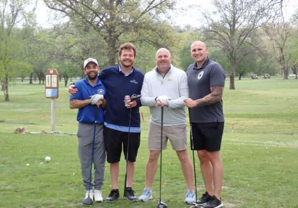 Four men posing on a golf course with clubs and drinks.